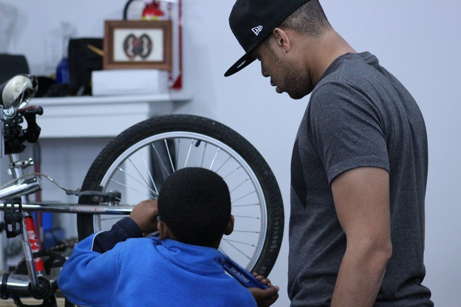 A child wearing a blue hoodie working on the back tire of a bicycle with an adult in a hat watching over 